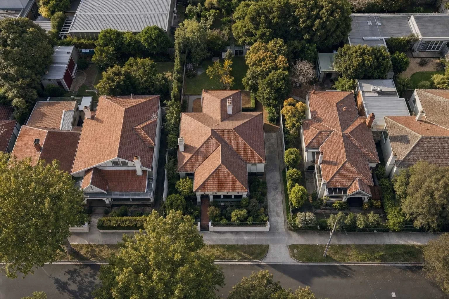 An aerial view of a suburban street with terracotta-roofed period homes, tree-lined gardens, and driveways leading to a central road