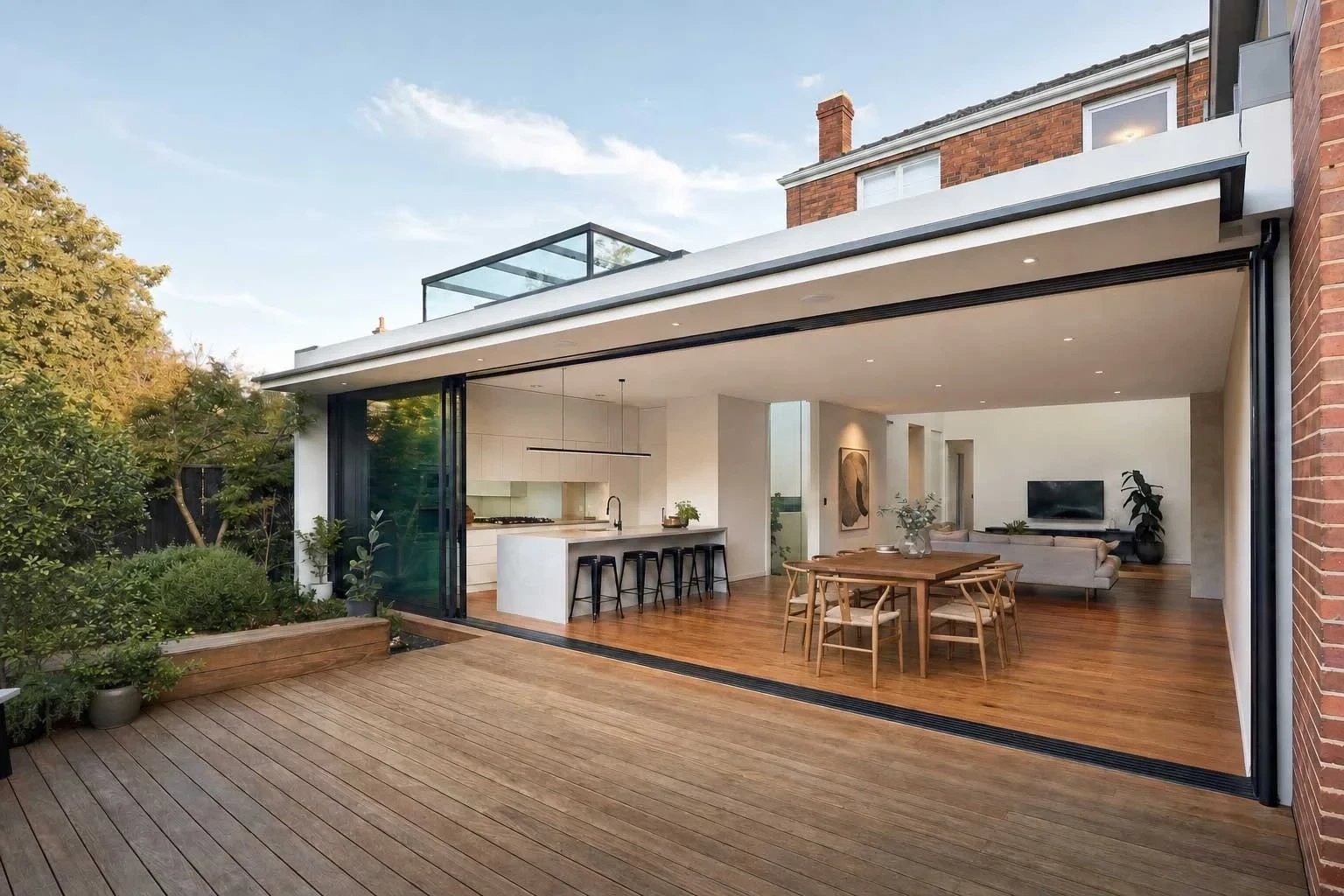 A period brick home's rear extension with black-framed sliding glass doors fully opened to an indoor-outdoor connection between a timber deck and an open-plan kitchen, dining and living space