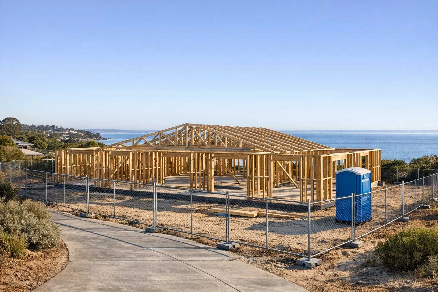 A timber house frame under construction on a coastal block with ocean views, surrounded by temporary security fencing and a portable toilet