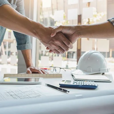 Two people shaking hands over a table with blueprints, a calculator, a pen, glasses, and a safety helmet, in an office environment.