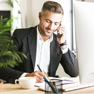 A man in a suit sits at a desk, talking on the phone, taking notes with a pen, near a computer monitor, with a coffee cup and some office supplies on the desk.
