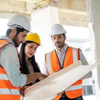 Three construction workers, two men and one woman, wearing safety helmets and orange vests, examining a large blueprint on a construction site.