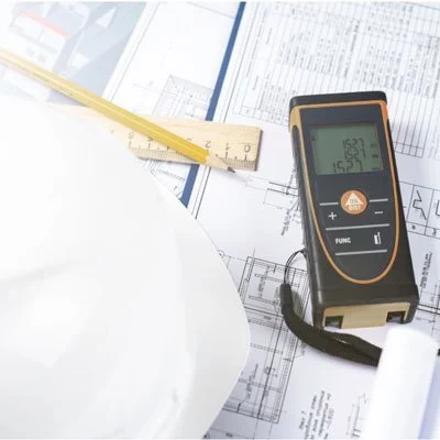 A laser distance measurer on architectural plans with a yellow measuring tape and a white helmet.