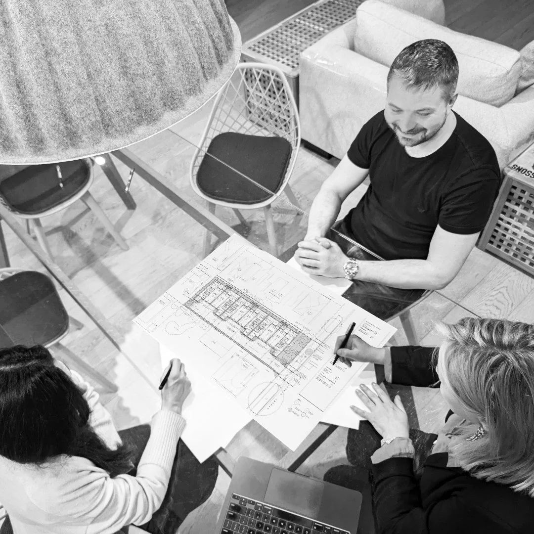 Overhead black-and-white photo of a design meeting: a man at a table smiling while two people opposite mark up an architectural floor plan, with a laptop on the table
