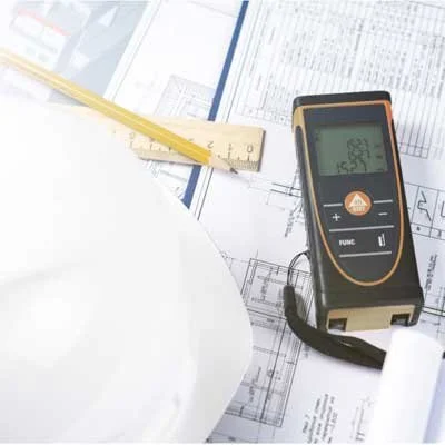 A laser distance meter on architectural blueprints, with a yellow ruler and a white plate nearby.