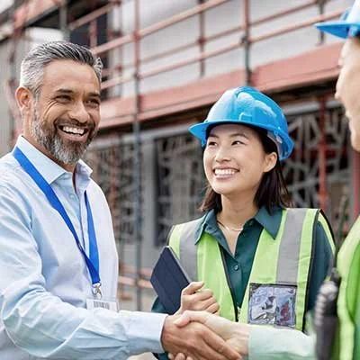 Two construction workers, a man and a woman, shaking hands at a construction site. The man has gray hair and a beard, wearing a white shirt and safety badge. The woman, wearing a blue hard hat and safety vest, is smiling.