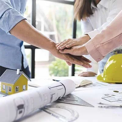 Three people placing their hands together in a teamwork gesture on a desk with blueprints and a small yellow house model.